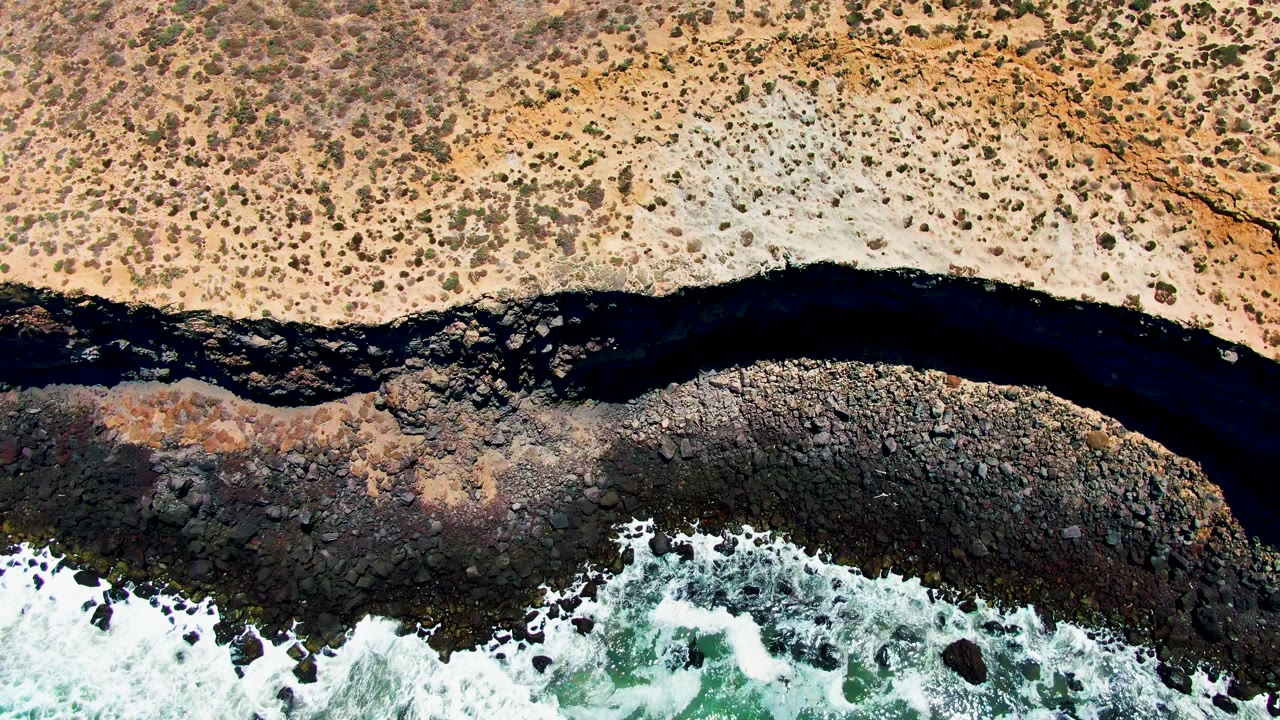 Aerial frame in San Quintin BC Mexico with the dark river band cutting between pale dry land and a black rocky shore as foamy waves push upward, with no visible people.