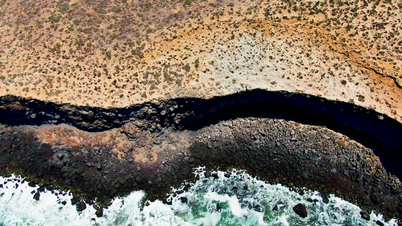 Top-down San Quintin BC Mexico coastline frame showing mottled tan ground, a winding dark watercourse, rounded dark rocks, and white ocean foam, with no visible people.