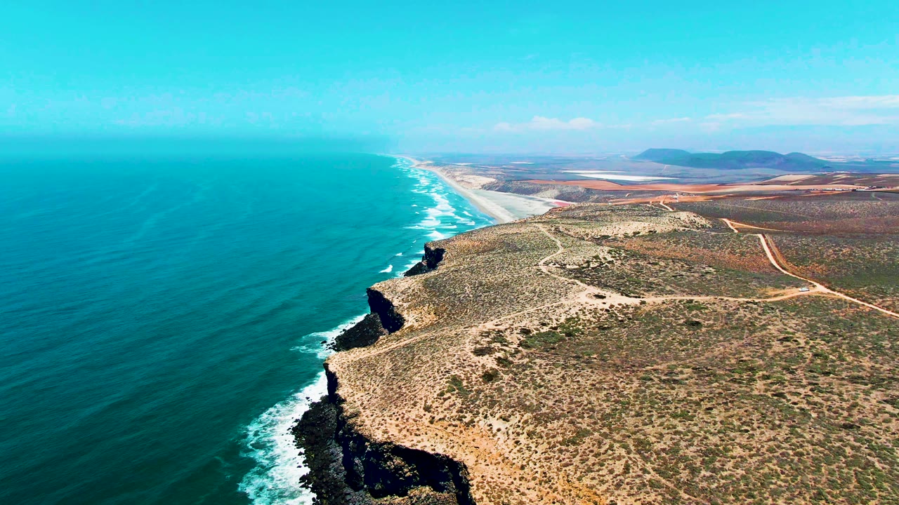 Aerial view of San Quintin BC Mexico one second into the clip, with the surf line extending farther up the beach, rocky headland in the lower right, dry patterned scrubland, distant hills, and no visible people.