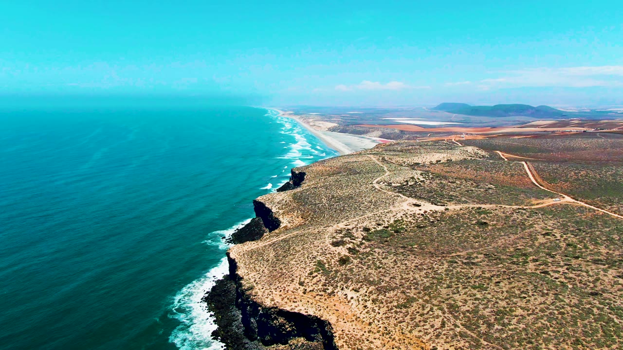 Aerial still from San Quintin BC Mexico showing turquoise water, white foam against the shoreline, a jagged dark cliff face, pale beach in the distance, scrub-covered coastal plain, and no visible people.
