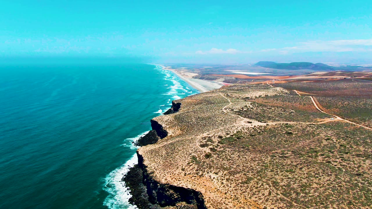 Mid-clip aerial frame over San Quintin BC Mexico with a rocky point in the lower right, ocean waves breaking along the left shoreline, sandy beach stretching into the distance, inland dirt tracks, and no visible people.