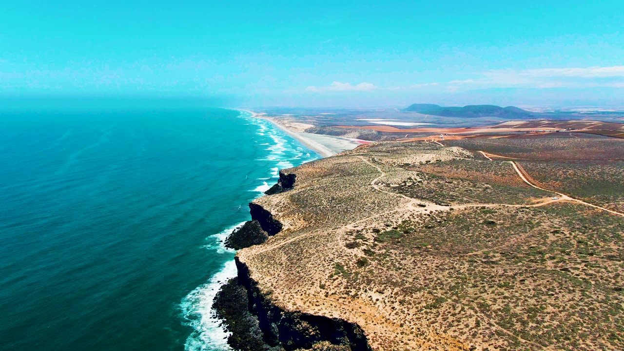 Aerial view of turquoise ocean, rocky cliffs, sandy beach, and arid coastal land.