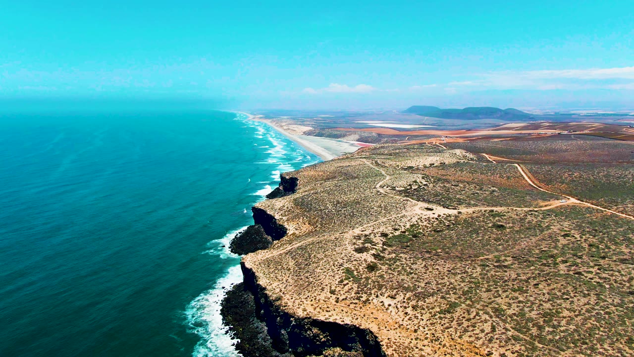 Aerial San Quintin BC Mexico coastline one second in, showing a long white surf line beside dark cliff faces, pale beach in the distance, dry inland terrain, and no visible people.
