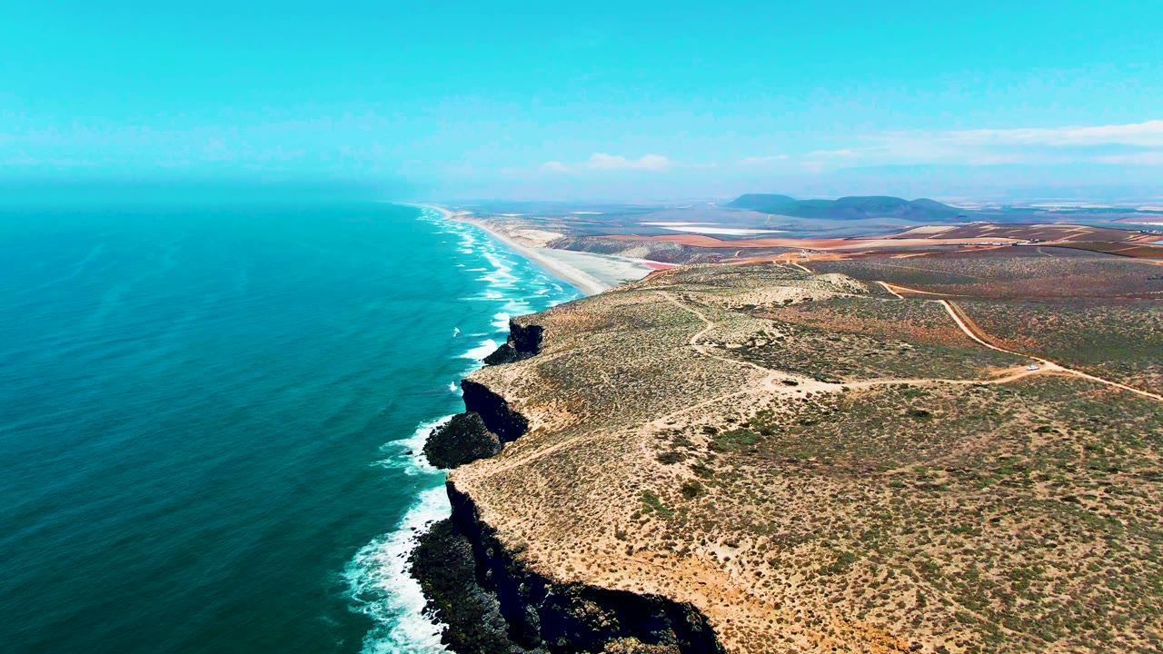 Aerial view over San Quintin BC Mexico with turquoise water occupying the left half, foamy waves at the cliff base, dry scrubland on the right, distant hills, and no visible people.
