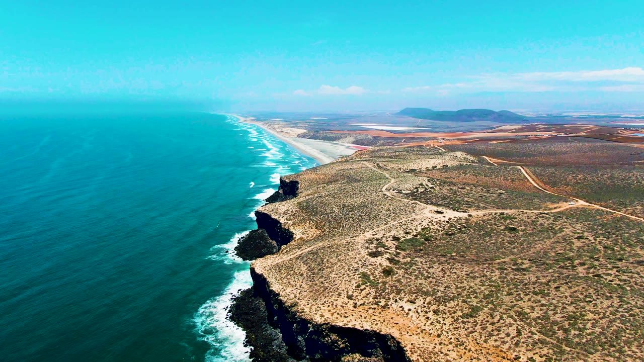 Mid-clip aerial frame of San Quintin BC Mexico shoreline, with dark cliff points jutting into turquoise ocean, white foam tracing the rocks, a long beach beyond, and no visible people.