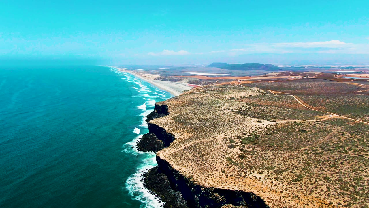 San Quintin BC Mexico coastline from above, with breaking white waves tracing the cliff base, tan brushland inland, long sandy shoreline in the distance, and no visible people.