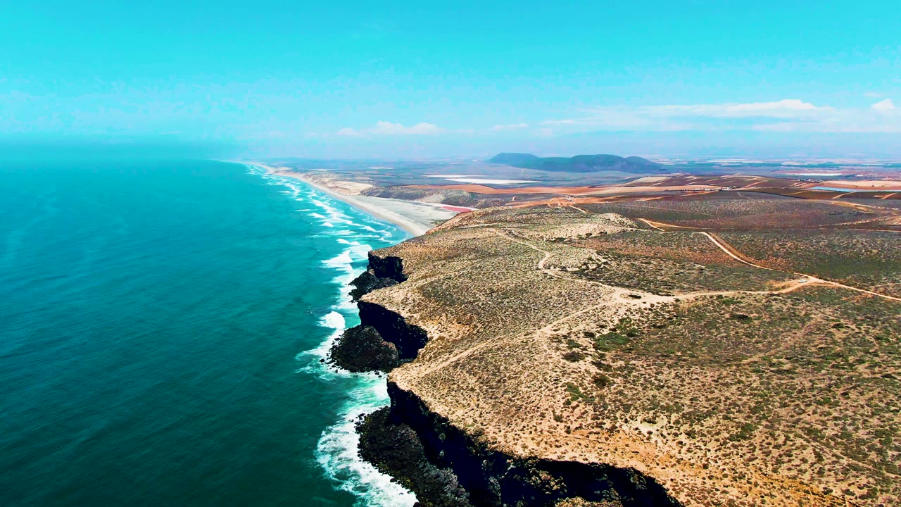 Mid-clip aerial frame over San Quintin BC Mexico, with surf curling beside black cliff sections, scrub-covered flats inland, pale beach farther up the coast, and no visible people.