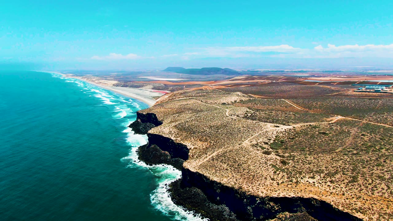 Opening aerial frame over San Quintin, Mexico, with blue ocean on the left, white surf on dark cliffs, dry scrubland inland, and no visible people.