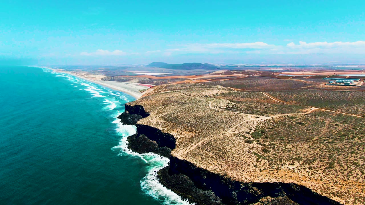 Aerial frame from San Quintin, Mexico, showing white foam wrapping the cliff base, a long beach stretching north, dry inland roads, and no visible people.