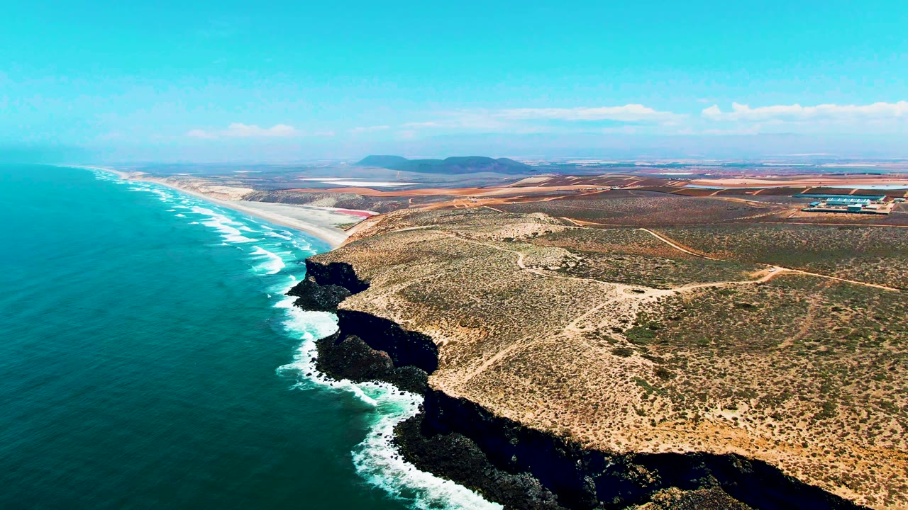 San Quintin, Mexico aerial view with the ocean occupying the left half, cliff shadows along the shore, pale sand beyond the rocks, sparse scrub inland, and no visible people.