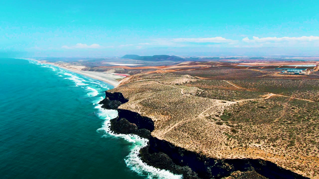 Aerial view of turquoise water beside rocky headlands and dry coastal roads.