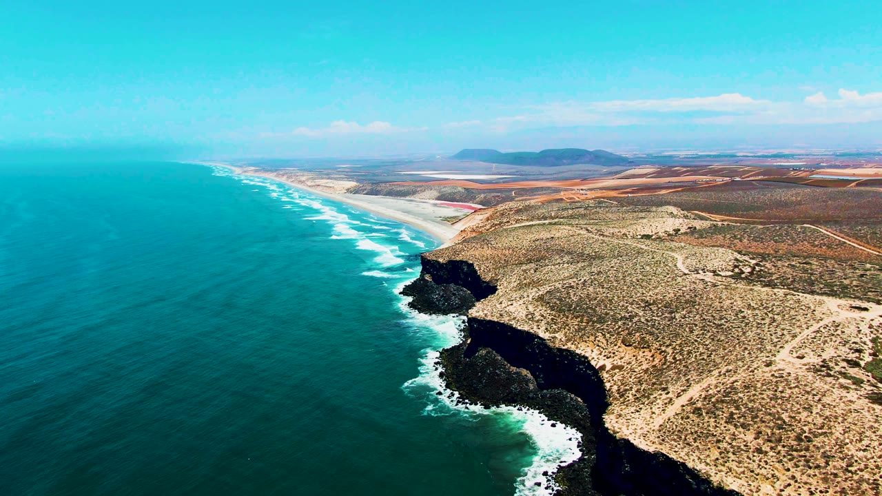 Aerial frame over San Quintin, Mexico, with turquoise ocean on the left, white surf around dark cliffs on the right, dry scrubland beyond, distant hills, and no visible people.