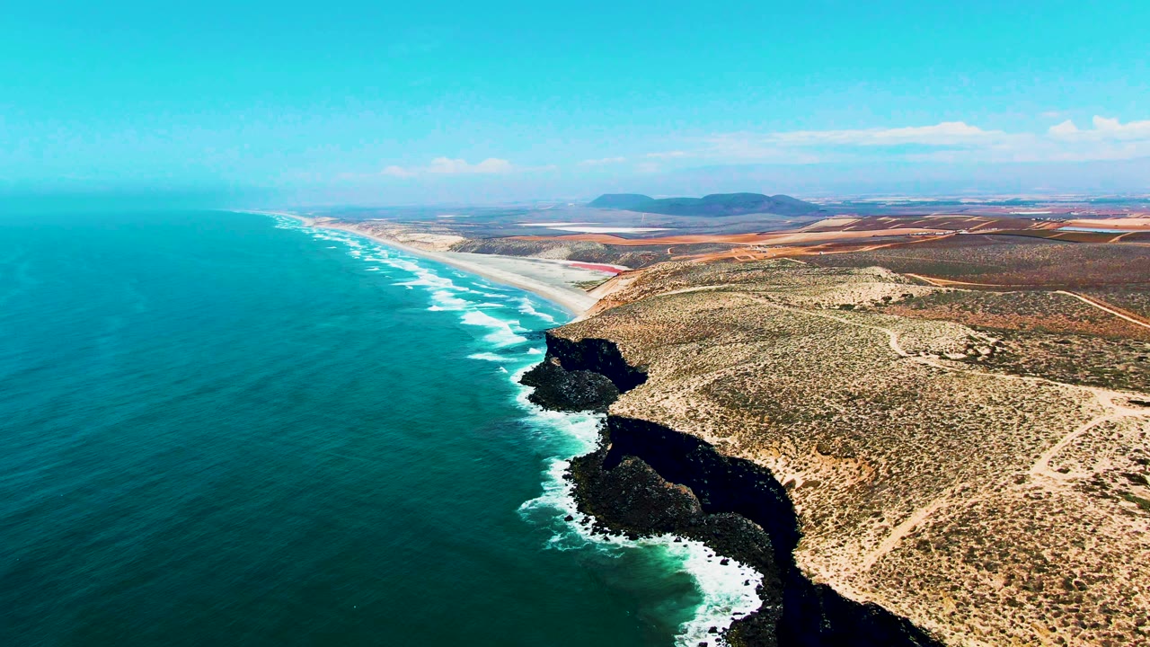 San Quintin, Mexico coastline seen from above, with foamy waves tracing the cliff base, open blue water stretching left, tan scrubland to the right, and no visible people.