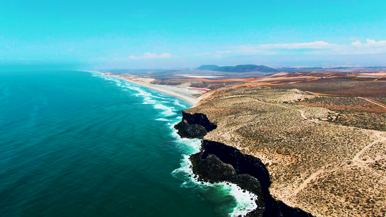 High drone frame over San Quintin, Mexico, showing a broad blue ocean, white breakers against black rock, pale dirt tracks across dry coastal ground, distant hills, and no visible people.