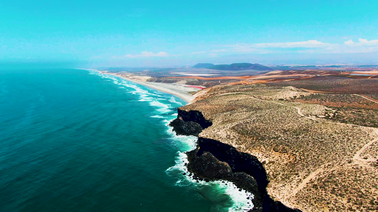 Drone view down a coastal cliff line with waves and arid land beside the sea.