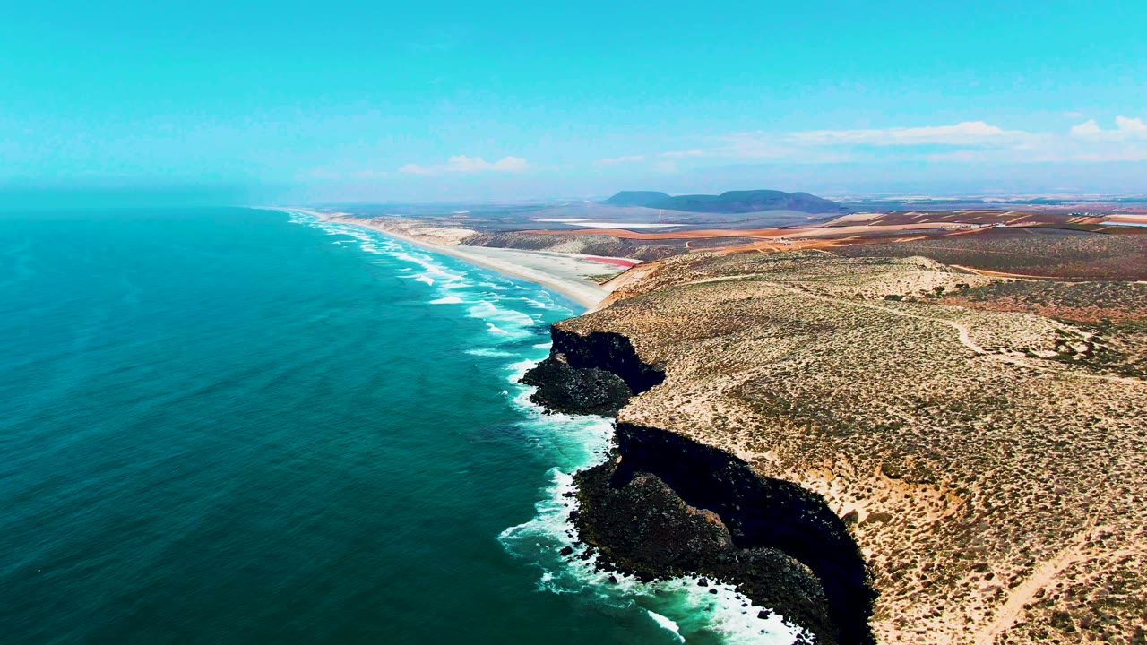 Aerial view of the San Quintin BC Mexico coast with turquoise ocean on the left, white surf along dark rocky cliffs, sandy beach in the distance, arid scrubland inland, and no visible people.