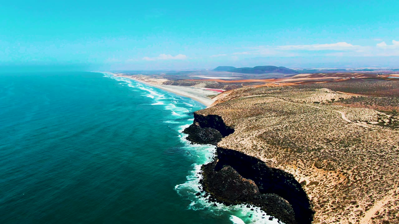 San Quintin BC Mexico shoreline seen from above, with a long sandy beach receding into the background, waves tracing the coast, dark cliff faces in the midground, and no visible people.