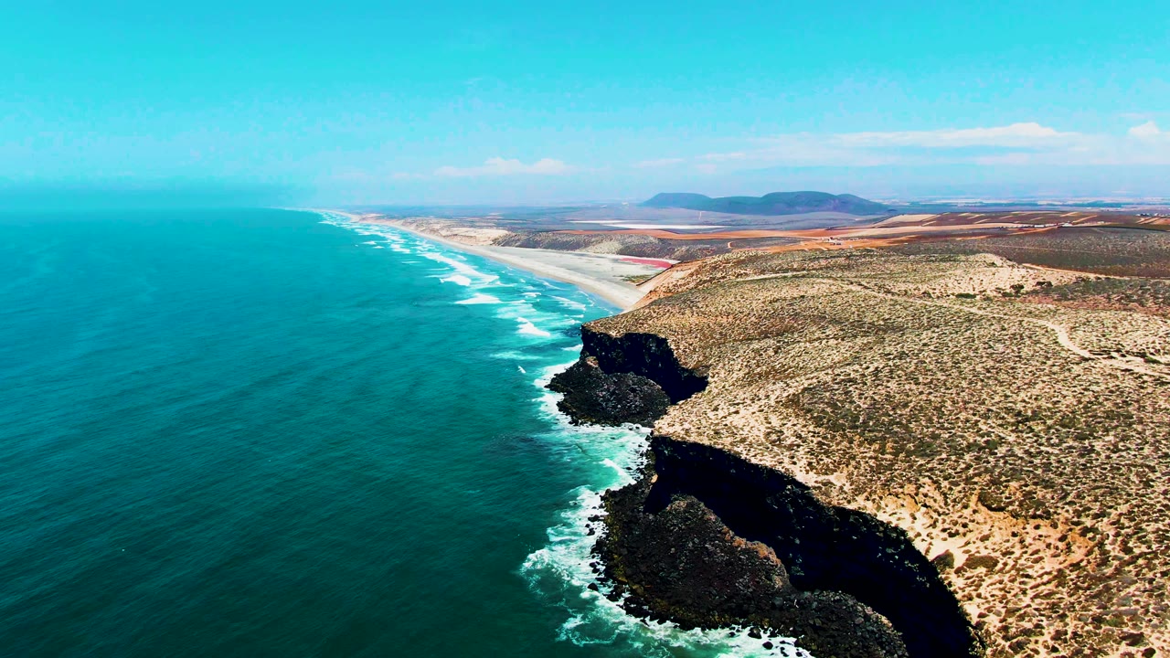 Aerial view of San Quintin BC Mexico coast with turquoise sea covering the left half, white wave lines along the shore, dark cliffs at lower right, sandy arid land inland, and no visible people.