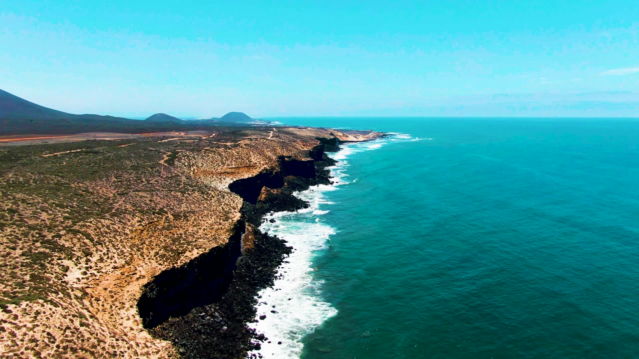 San Quintin, BC, Mexico coastline from above, with dark cliff shadows in the foreground, surf foam tracing the rocks, open turquoise water to the right, and no visible people.