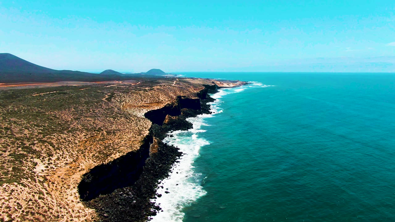 Mid-clip aerial view over San Quintin, BC, Mexico showing the rocky headland receding into the distance, white surf bands along the cliff base, blue-green ocean, and no visible people.