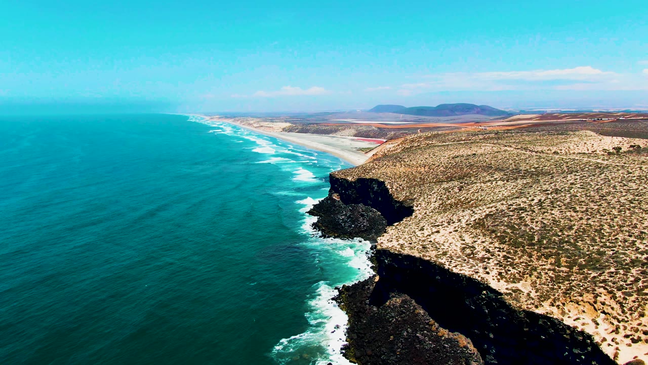Aerial drone view of the rocky Pacific coastline near San Quintin, Baja California, Mexico, showing a dark volcanic headland, turquoise ocean, white surf foam, dry desert terrain, and distant mountains. No people visible.
