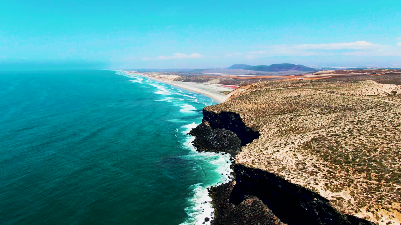 Wide aerial drone view of the San Quintin, Baja California coast, with dark volcanic headland, turquoise Pacific Ocean, white wave foam, dry scrubland terrain, and a long beach extending toward the mountain horizon. No people visible.