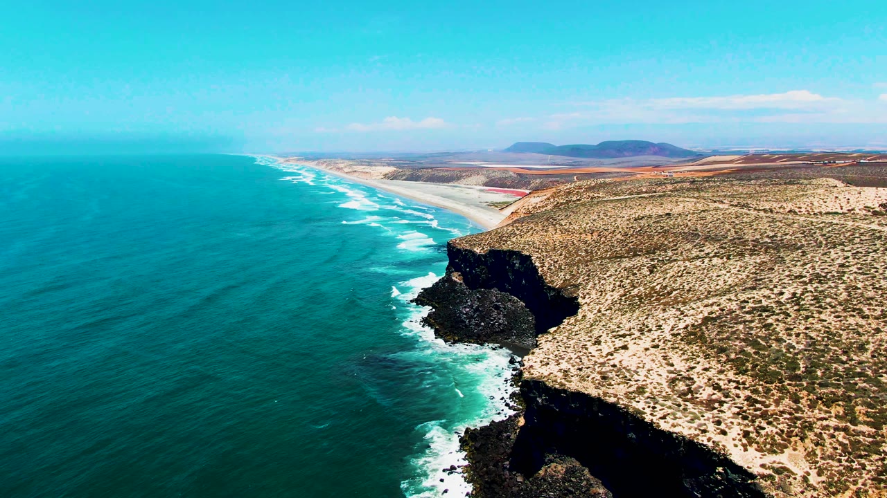 Aerial drone shot from a forward or lower position over the San Quintin, Baja California headland, with the dark cliff face filling more of the foreground, turquoise Pacific, white surf foam, and beach stretching into the distance. No people visible.