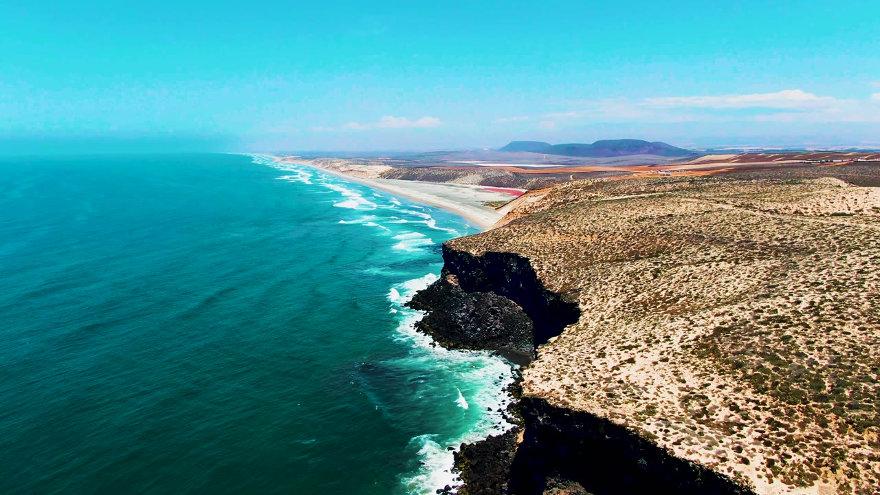 Aerial drone shot over the San Quintin, Baja California rocky headland, with the dark volcanic cliff prominent in the foreground, turquoise ocean, white surf, dry scrubland, and the distant coastline. No people visible.