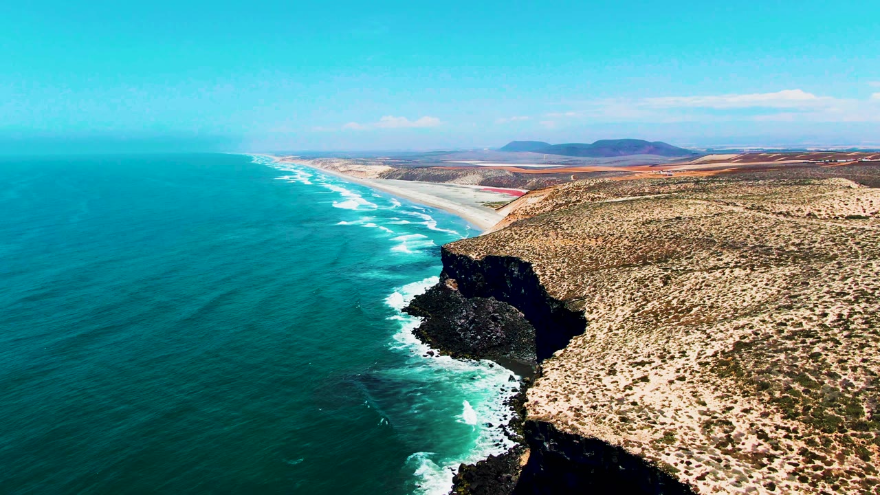 Final aerial drone frame over the San Quintin, Baja California coast, with the dark rocky headland in close foreground, turquoise Pacific Ocean, white surf, dry desert terrain, and mountains on the distant horizon. No people visible.