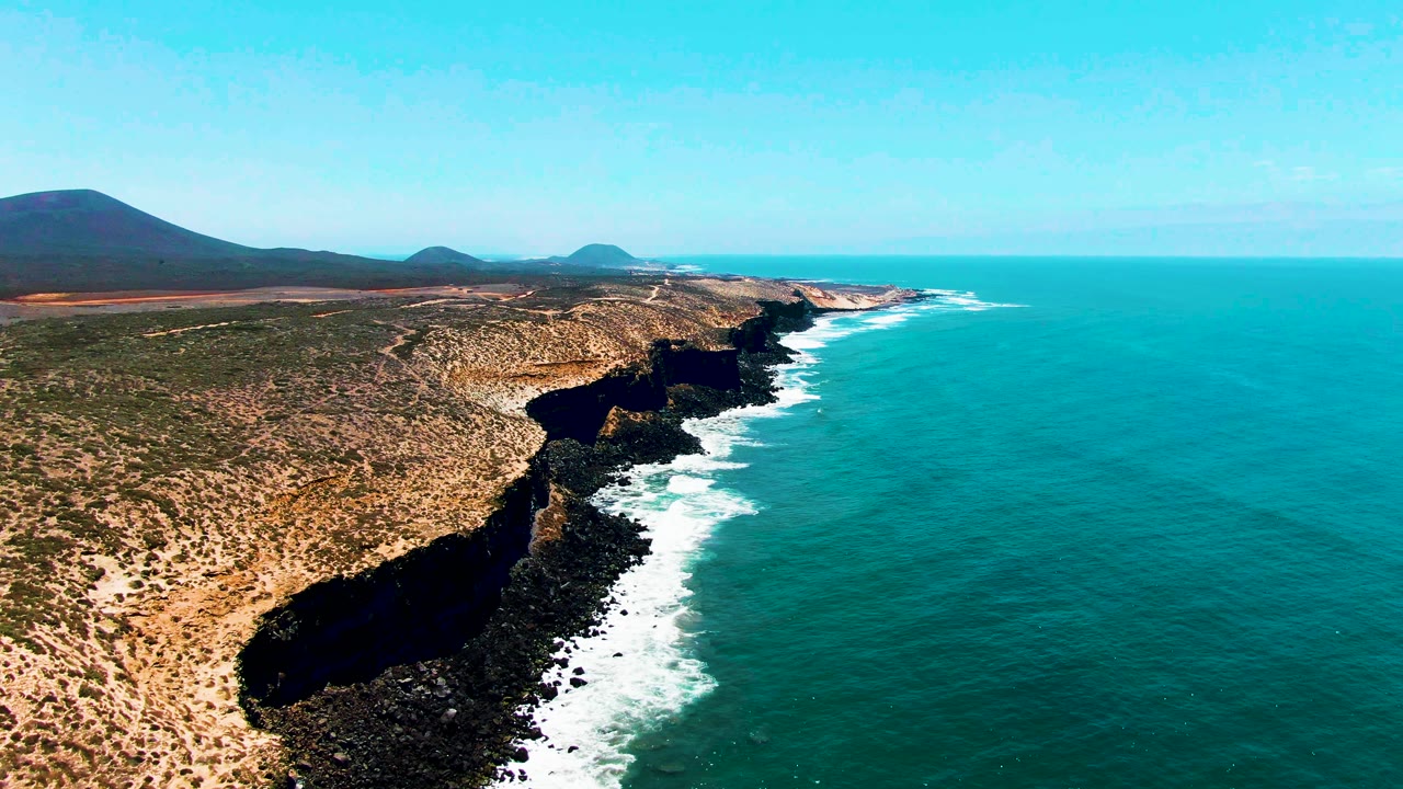 Aerial view over San Quintin BC Mexico showing dry rocky cliffs on the left, turquoise ocean on the right, white surf along the shore, distant hills, and no visible people.
