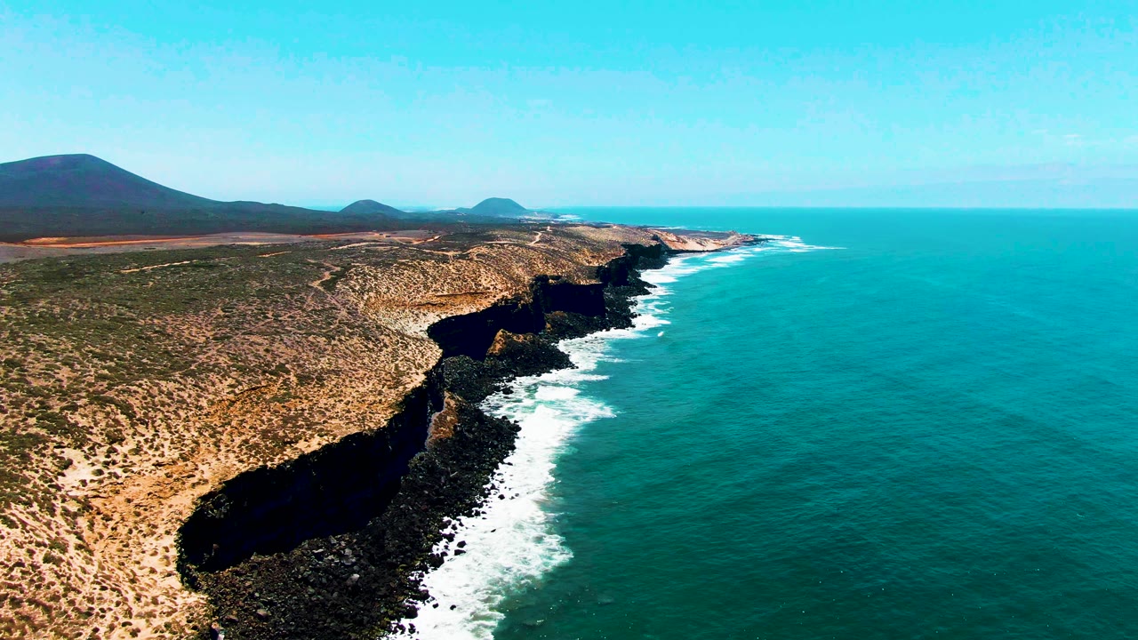 High aerial frame of San Quintin BC Mexico with white waves tracing the dark cliff base, scrubby dry land inland, blue ocean offshore, distant hills, and no visible people.
