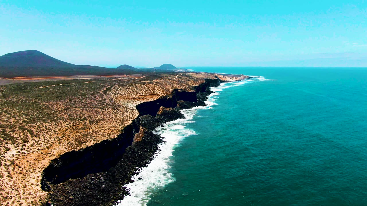 Aerial coastline with surf washing against rugged cliff shadows.