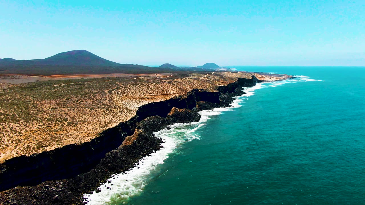 Aerial view of rocky coastal cliffs, turquoise ocean, white surf, dry land, and distant mountains.