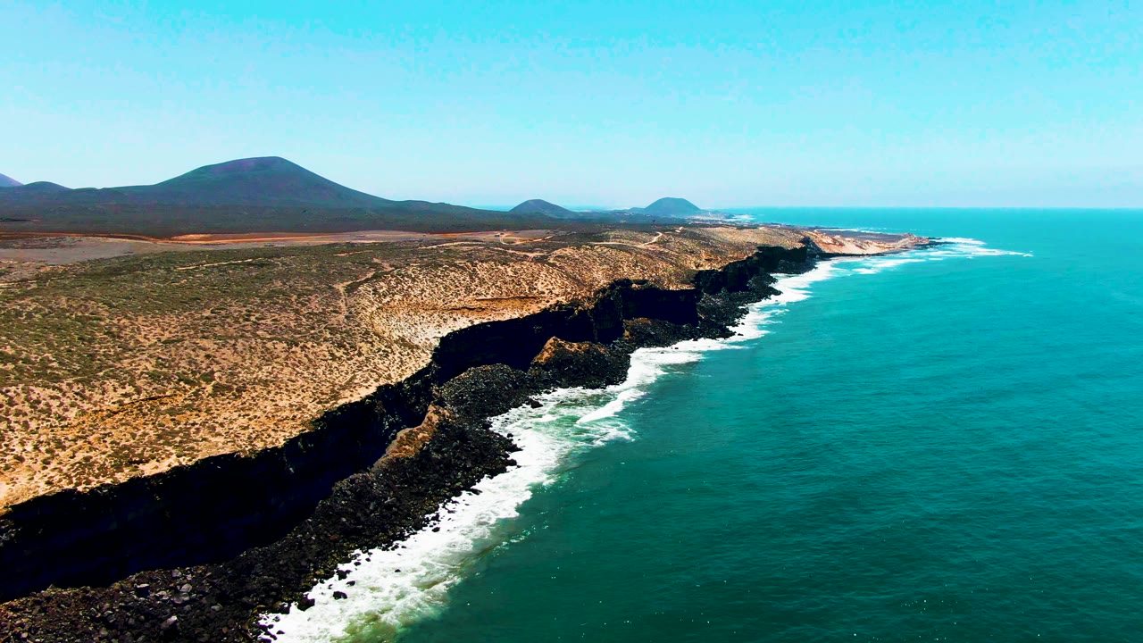 San Quintin, Mexico coastline seen from above with blue-green water, foamy wave lines against dark rocks, arid land running inland, low mountains behind it, and no visible people.