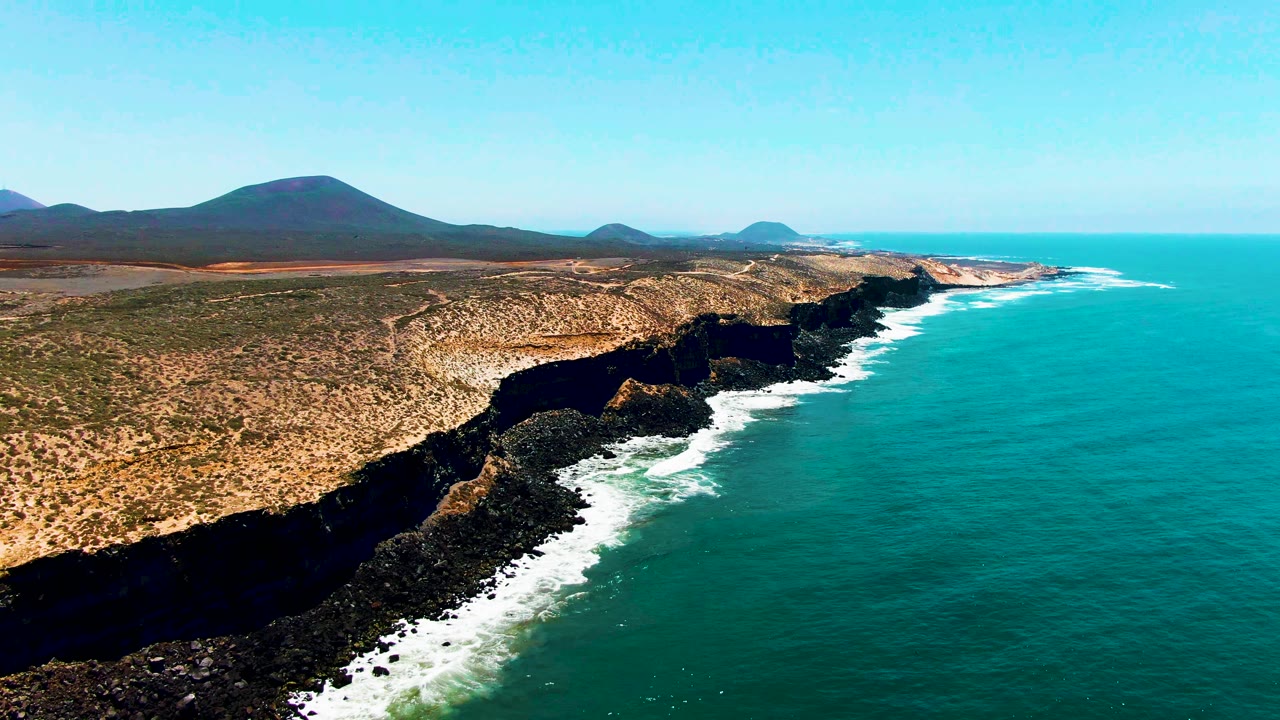 Wide aerial frame over San Quintin, Mexico showing turquoise sea to the right, white surf along the cliff base, dry tan terrain inland, several distant mountain forms, and no visible people.