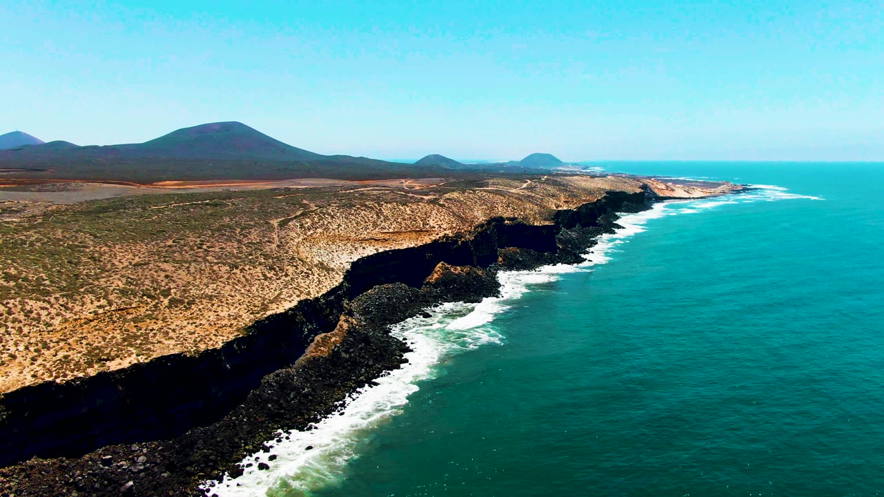San Quintin, Mexico aerial view with jagged dark cliffs cutting between turquoise ocean and scrub-covered dry land, white water collecting along the rocks, and no visible people.