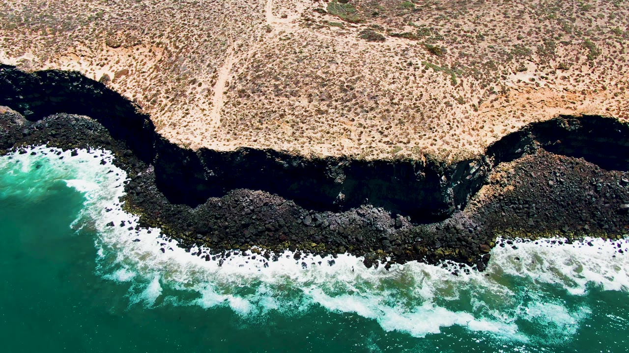 Aerial view in San Quintin, Mexico of turquoise water breaking against a dark rocky ledge beneath dry tan coastal scrub, with no visible people.