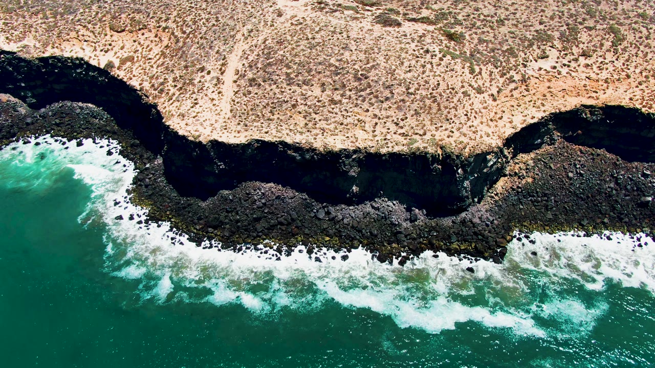 Aerial frame from San Quintin, Mexico showing white foam spreading farther along a curved dark rock shoreline below dry coastal terrain, with no visible people.