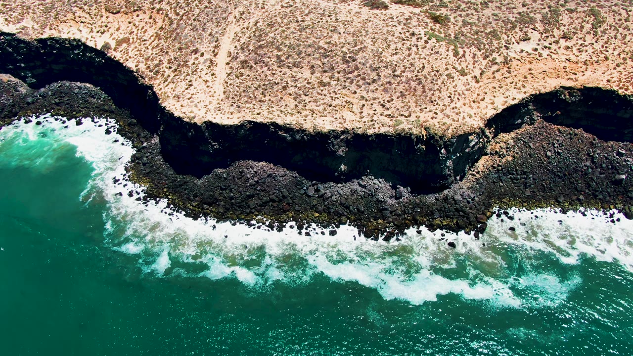 Top-down San Quintin, Mexico view with a fresh band of white surf striking the jagged dark coastline and beige scrubland holding static above it, with no visible people.