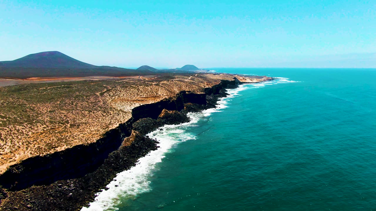 Opening aerial frame over San Quintin BC, Mexico with turquoise ocean to the right, white surf against dark rocky cliffs, arid land to the left, distant hills on the horizon, and no visible people.