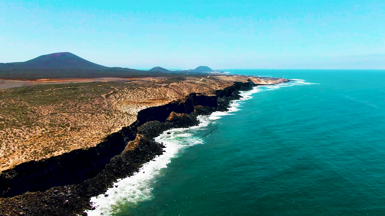 Mid-clip aerial over San Quintin BC, Mexico showing dark rocky shoreline, white wave foam, broad turquoise water, tan arid ground, distant low hills, and no visible people.