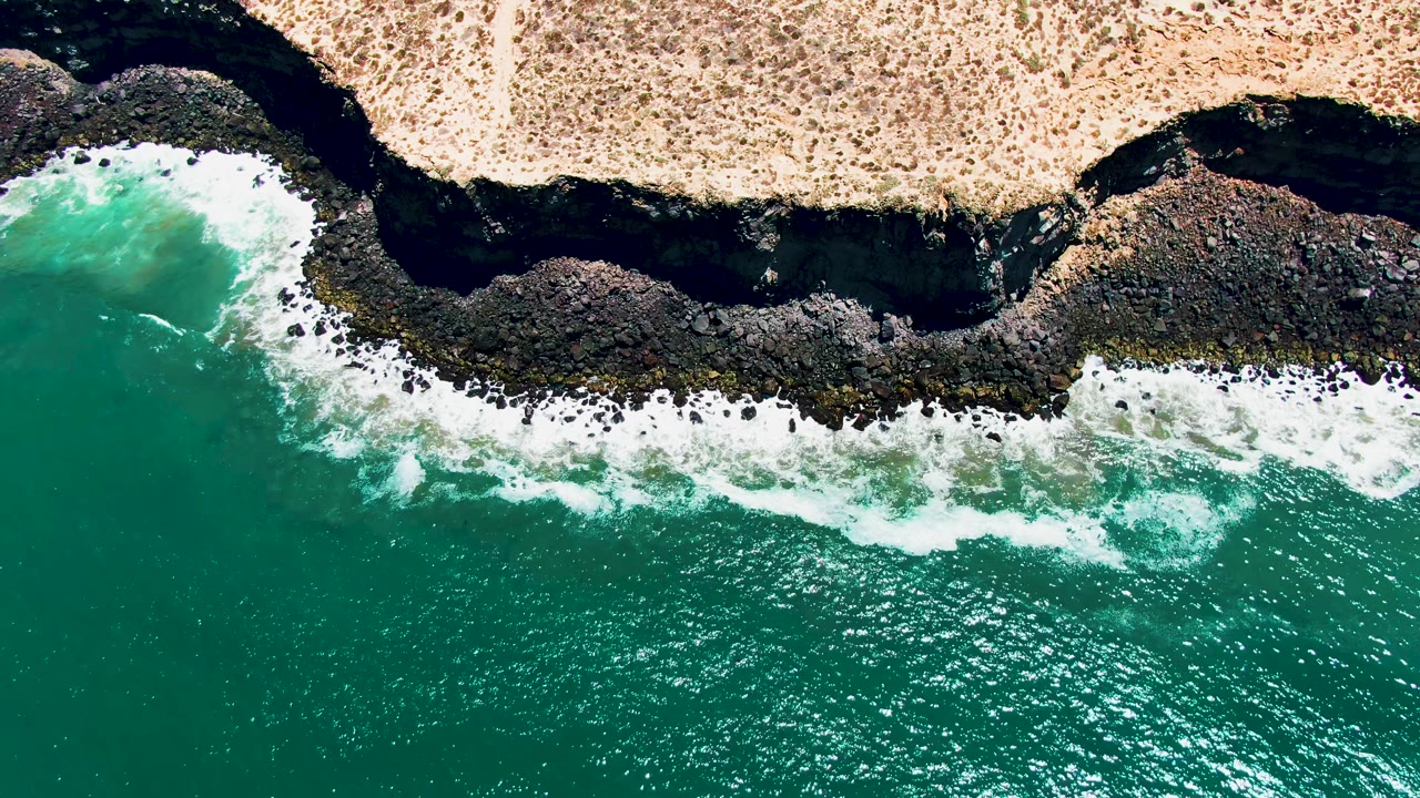 Overhead aerial frame from San Quintin BC Mexico showing turquoise water, white surf, dark shoreline rocks, dry sandy land, and no visible people.