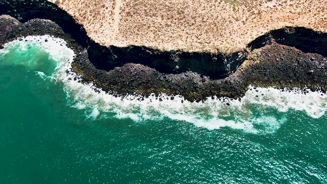 Aerial view over San Quintin BC Mexico with white wave foam widening along the rocks, turquoise ocean below, dry land above, and no visible people.