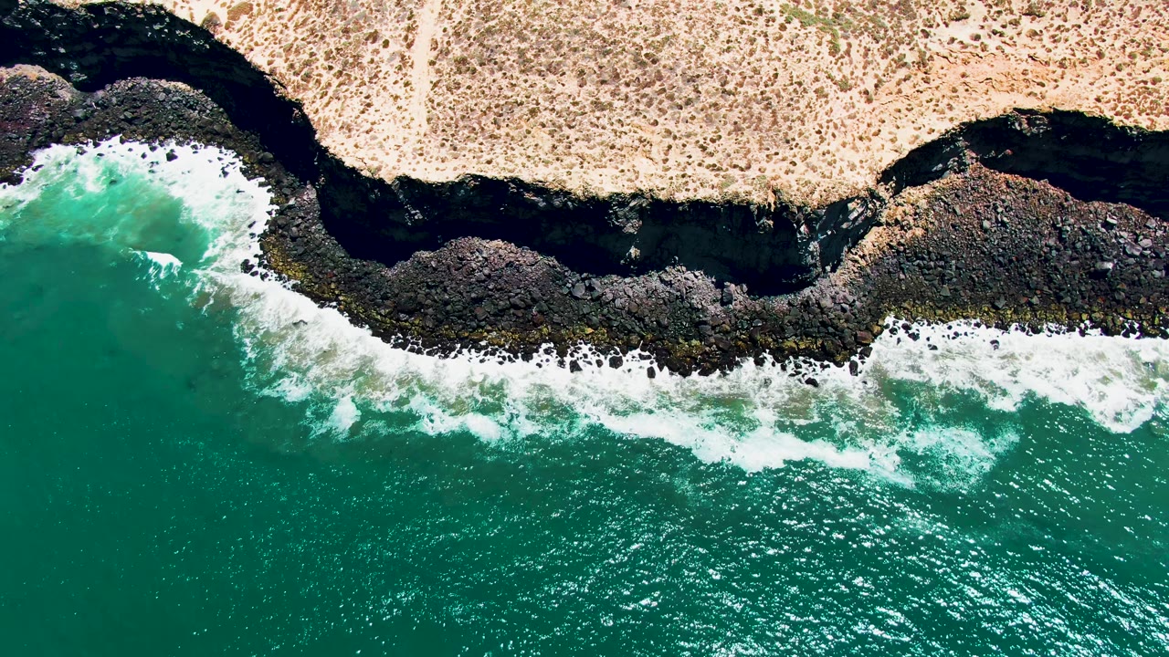 Top-down San Quintin BC Mexico coastline frame with a bright foam line curling into a dark rocky indentation, sandy dry land above, and no visible people.