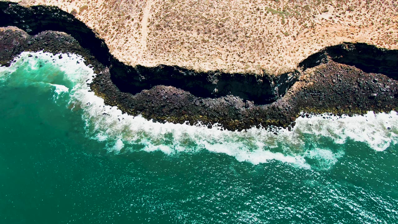 Aerial coastline view with foam breaking across black rocks and turquoise water.