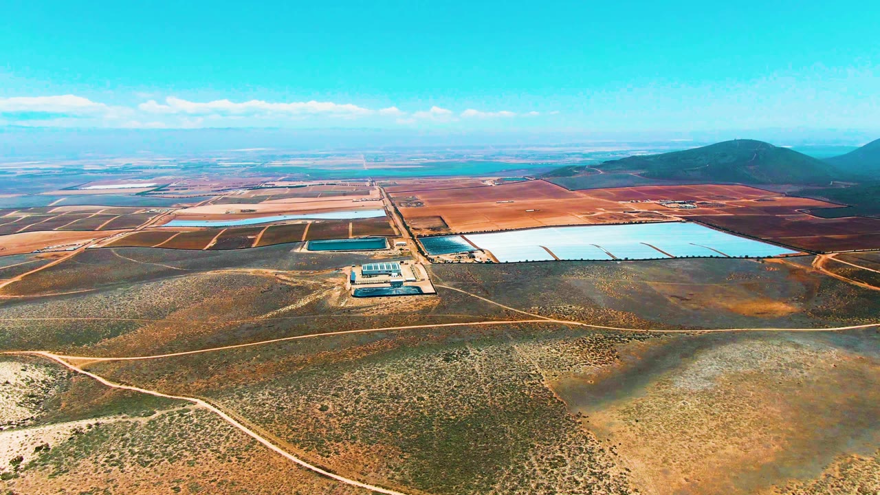 Aerial view over San Quintin BC Mexico showing pale reservoir basins, smaller dark ponds, dry scrubland, dirt roads, distant hills, blue sky, and no visible people at the start of the clip.