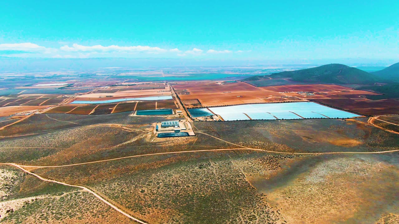 High aerial view in San Quintin BC Mexico with rectangular pale reservoirs, dark holding ponds, tan dirt tracks, scrub-covered foreground, distant fields, hills, and no visible people.