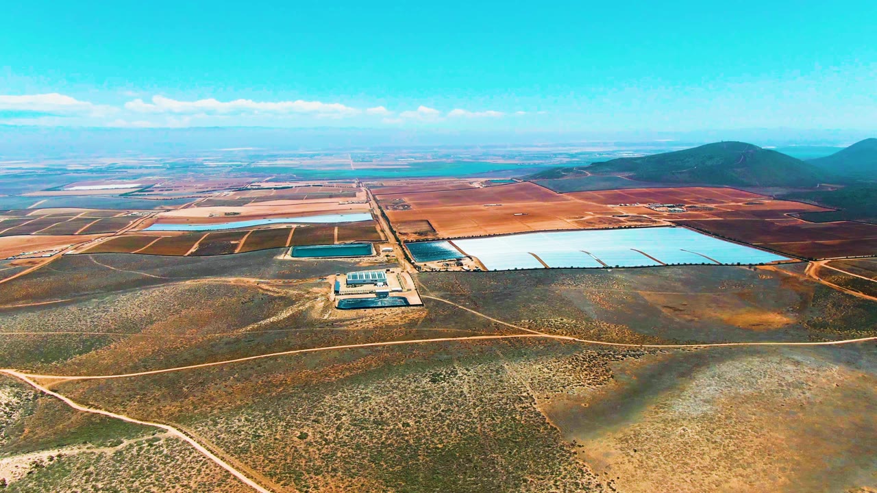 Drone-like aerial frame over San Quintin BC Mexico showing bright pale water basins, a small built structure near the ponds, brown arid terrain, low vegetation, hills on the horizon, and no visible people.