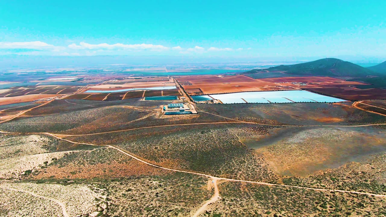 Wide aerial frame over San Quintin BC Mexico showing dry scrubland crossed by dirt roads, rectangular industrial water ponds in the distance, low hills on the horizon, and no visible people.