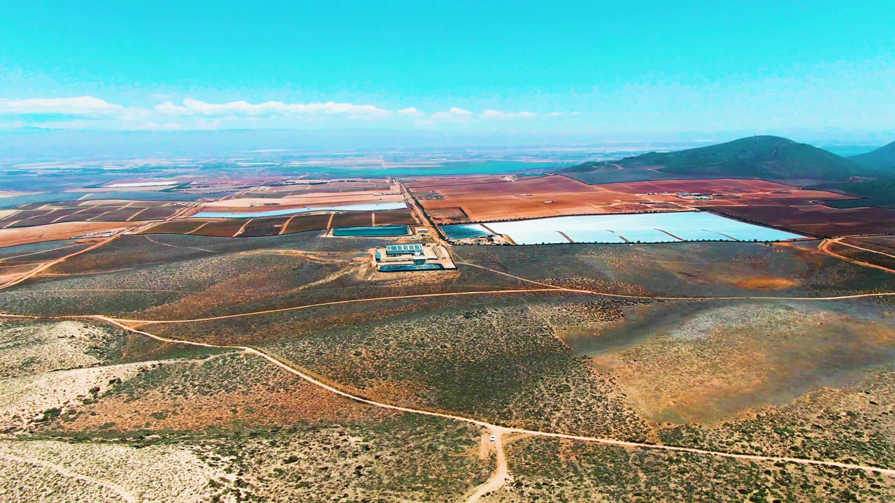 Drone-height view across dry terrain toward rectangular ponds and low hills under clear sky.